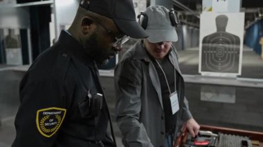 Afrika kökenli Amerikalı polis memuruna eğitim veren, çeşitli silah çeşitlerini gösteren ve özelliklerini açıklayan beyaz erkek atış antrenörünün orta boy yakın çekimleri.