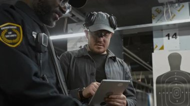 Afrika kökenli Amerikalı erkek stajyer güvenlik ajanının atış poligonunda silah kullanmayı öğrenirken tabletli Kafkasyalı eğitmen de izliyor, düzeltiyor ve listeyi dolduruyor.