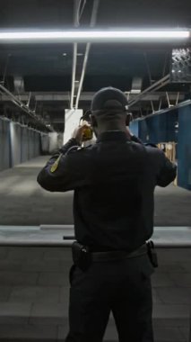 Dikey görüntü, üniformalı Afro-Amerikan polis memurunun atış mesafesinde durup, kulaklık takıp, uzaktan hedefe silah doğrultup keskin nişancılık becerilerini denerken görüntüsü.