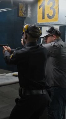 Beyaz erkek eğitmenin, Afrika kökenli Amerikalı stajyer polis memuruna doğru atış pozisyonu ve kavramayı öğretirken, aynı zamanda kapalı alanda tabanca kullanma becerileri konusunda eğitim verdiği orta boy dikey görüntü.