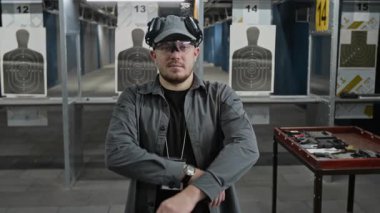 Genç beyaz erkek atış poligonu eğitmeninin orta boy portresi ya da iş yerindeki iş sahibi tabancalı, cephaneli ve mermili masaların yanında kameraya özgüvenle bakıyor.