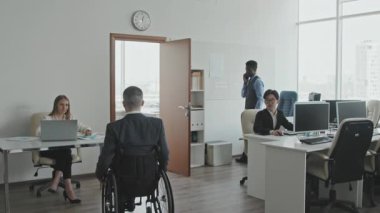 Tekerlekli sandalyedeki erkek yöneticinin iş yerindeki çalışanlar tarafından karşılandığı yavaş çekim, sonra da kadın kişisel asistanıyla birlikte belgelerle ofisine girmesi.