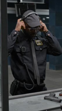 Orta dikey olarak kendine güvenen Afro-Amerikan güvenlik görevlisi veya federal ajan atış poligonunda duruyor, kulaklık takıyor, hedefe silah doğrultuyor, yeterlilik sınavına hazırlanıyor.