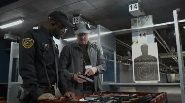 Orta yaşlı, beyaz bir erkek, eğitim gören siyah üniformalı Afro-Amerikan polisine nişancılık kursu sırasında şarjörleri tabancayla doldurmayı öğretiyor.