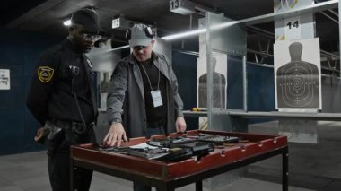 Orta boy Afro-Amerikan polis memuru ve beyaz erkek şut koçu. Galeride ekipmanla birlikte masanın yanında duruyor. Çeşitli tipte tabanca ve mühimmattan bahsediyorlar.