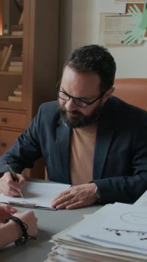 Gözlüklü beyaz erkek okul psikoloğunun müşteri dinlerken ve modern rahat ofisteki masada notlar yazarken çekilmiş dikey bir resmi.