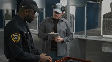 Siyahi üniformalı Afrika kökenli Amerikalı erkek polis, rozeti ve kılıfı ile atış poligonu sahibine tabletle konuşuyor, iş yerinde çeşitli tipte tabanca kullanma deneyimini paylaşıyor.