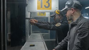 Orta halli beyaz erkek eğitmen, stajyer Afro-Amerikan polis memuruna atış poligonunda çalışırken nişan almasını, silah tutmasını, nişan almasını ve tetiği çekmesini öğretiyor.