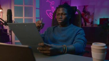 Orta karar, düşünceli, odaklanmış Afrikalı Amerikalı erkek podcaster ya da örgülü saçlı sivil gazeteci masasında oturuyor, röportaj planı üzerinde çalışıyor ya da sesli mesaj üzerinde çalışıyor.