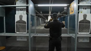 Siyahi bir polis ya da güvenlik görevlisinin kapalı alanda durup, kulaklık takıp, ateşli silah kuşanıp, gövde hedefine nişan alıp,