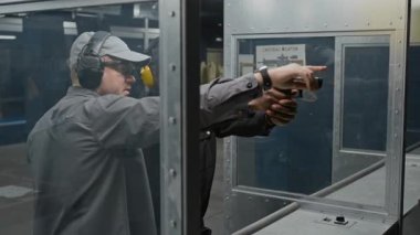 Orta yaşlı, beyaz erkek atış eğitmeni stajyer siyah polis memuruna üniforma ve kulaklık takmayı öğretiyor. Silah namlusuna nişan alma tekniği ve silah namlusuna nişan alıyor.