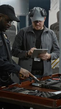 Siyah üniformalı Afrikalı Amerikalı stajyer güvenlik görevlisinin dikey orta sahası mühimmatı tabancaya yükleme çalışması tablet izleme ve performans kontrol listesiyle Kafkas koçu