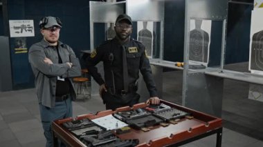 Afrika kökenli Amerikalı erkek polis memuru ve beyaz eğitmenin orta boy portresi silah, cephane ve mermilerle birlikte kameraya bakarak yan yana duruyor.