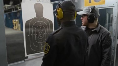 Kulaklık takan ve koruyucu gözlük takan erkek atış eğitmeninin orta dereceli kavisi ilk eğitim sırasında siyah üniformalı stajyer polis memuruna siluet hedefini gösteriyor.