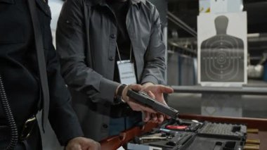 Kafkas erkek atış eğitmeninin orta eğik pozisyonu, siyah erkek güvenlik görevlisinin şarjörleri silaha nasıl dolduracağını gösteriyor, eğitim sırasında yapıcı özellikleri ve hareketleri anlatıyor.