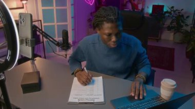 Genç Afrikalı Amerikalı erkek blogcunun laptoptaki popüler sosyal medya eğilimlerini araştırırken mor neon ev stüdyosunda içerik planı yazarken orta açıdan çekilmiş bir fotoğrafı.