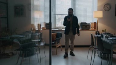 Orta yaşlı, sakallı, şık kıyafetler ve gözlüklü bir adamın modern ofiste cam bölme duvarlarıyla dolaşması ve telefonda sesli mesaj kaydetmesi.