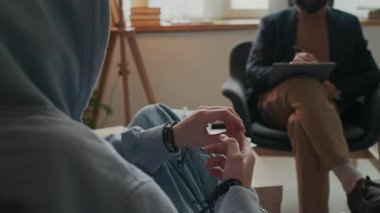 Psikoterapi gören depresif bir gencin omuz üstü fotoğrafı, sinir bozucu bir şekilde parmaklarına dokunarak ofisteki ruhsal sağlık veya okul baskısı sorunlarını tartışıyor.