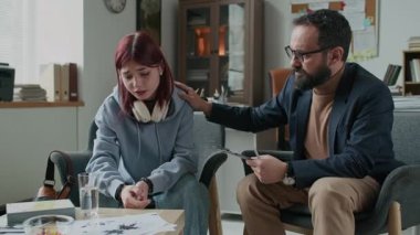 Kafkasyalı erkek psikoterapistin üzgün genç kızı dinleyip omzunu sıvazlayarak güvenini tazelediği ve terapi seansında duygusal destek verdiği orta boy bir fotoğraf.