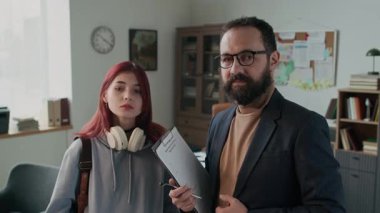 Sakallı beyaz bir erkek psikoloğun portresi şık giyinmiş ve gözlüklü üzgün kızıl saçlı, kulaklıklı, bol elbiseli, kameraya bakan bir kızla poz veriyor.