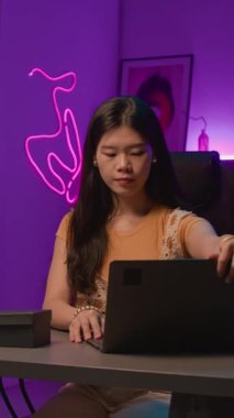 Genç Asyalı kadın nüfuzlunun stüdyoda masada otururken, dizüstü bilgisayarın kapağını açarken, podcast ya da canlı yayın için mikrofonu ayarlarken orta dikey görüntüsü.