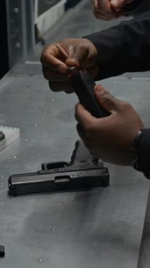 Siyahi erkek polis memurunun ellerinin ve yüzünün dikey yakın çekimi. Kurşunları şarjöre doldurup yarı otomatik silaha yerleştiriyor. Hedefe ateş ediyor ve nişan alıyor.