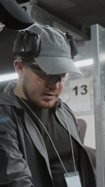 Profesyonel Kafkas erkek atış eğitmeninin dikey ortamı antrenmandan önce çeşitli tabancalarla masada duruyor ve ekran dışı müşterilere danışmanlık yapıyor.