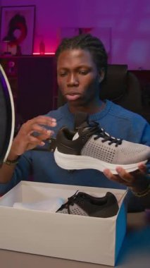 Süslü saçlı, stüdyo masasında oturan Afro-Amerikalı bir erkeğin orta boy dikey görüntüsü. Sosyal medyada veya çevrimiçi mağazada izleyicilere yeni spor ayakkabıları incelemesi