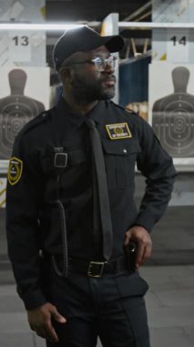 Atış poligonunda durup, atış talimi yapan kendinden emin Afrikalı Amerikalı erkek polis memurunun orta boy dikey portresi. Kabinler ve hedefler önünde kameraya bakıyor.