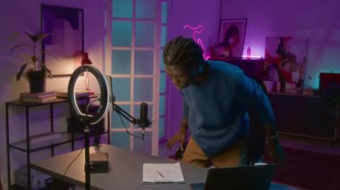 Orta boy Afro-Amerikan erkek etkileyicinin halka ışığı ve akıllı telefonu tripoda kurarken, kamerayı açarken, dizüstü bilgisayarla masaya otururken, yayına hazırlanırken,