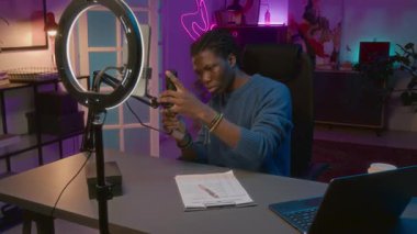 Meşgul genç Afrikalı Amerikalı erkek blogcu kabloyu mikrofona takıyor, sesi kontrol etmek için tıklıyor, sonra dizüstü bilgisayarı açıyor ve içerik planı için sosyal medya eğilimlerini araştırıyor.