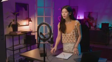 Çinli kadın nüfuzcunun tripoda halka ışığı açması, akıllı telefondan kamerayı açması, masaya oturup gülümsemesi, sosyal medyada canlı yayına başlamaya hazırlanması.