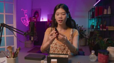 Genç Koreli kadın güzellik blogcusu stüdyoda canlı yayında mikrofonun önünde kozmetiklerle dolu kutuları açıyor, yüz serumunu çıkarıyor ve cilde damlalar uyguluyor.