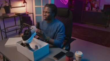 Sosyal medya yayını sırasında spor ayakkabılarının yeni modelinin reklamını yapan kendinden emin Afrikalı Amerikalı erkek içerik üreticisinin orta açı çekimi, modaya uygun tasarım ve kaliteli malzemelere övgüde bulundu