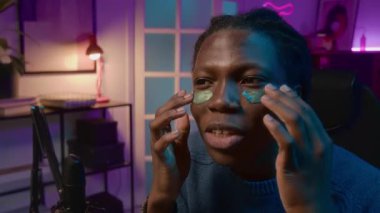 Sosyal medyada ya da internet sitesinde canlı yayın sırasında nemlendirici göz bantları deneyen neşeli Afrikalı Amerikalı erkek blogcunun kapanışı, kozmetik ürün faydalarından bahsediyor