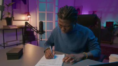 Genç siyah erkek podcaster 'ın örgü örgülü saçlarıyla orta kavisli görüntüsü masada oturuyor, dizüstü bilgisayardaki haberleri araştırırken aynı zamanda ev yayın stüdyosundaki planlanmış podcast için içerik planı yazıyor.