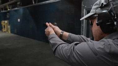 Gri beyzbol şapkalı, koruyucu kulaklıklı, şarjörü dolduran, kapalı alanda çalışırken gövde hedefine ateş eden genç adamın yüzünün ve ellerinin yan görüntüsü.