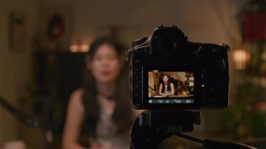 Tripodlu profesyonel video kamera ekranının kapanışı. Genç Çinli kadın görüş lideri canlı yayında podcast yapıyor. Kayıt stüdyosunda masa başında oturuyor.