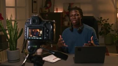 Orta ölçekli duygusal, canlı Afro-Amerikan gen z erkek podcaster kameranın önünde üç ayaklı sandalyede oturuyor. Ev kayıt stüdyosunda, trend konular ve sorunlar hakkında konuşuyor.