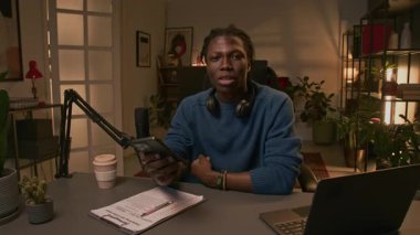 Orta boy, gülümseyen siyah erkek blogcu. Mikrofon ve dizüstü bilgisayarla masada oturmuş abone sorularını okuyor ve görüşlerini bildiriyor.