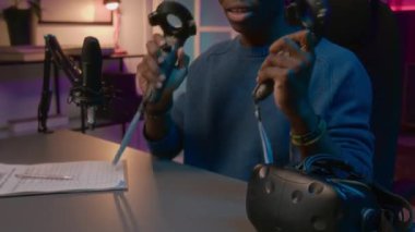 Heyecanlı genç Afrikalı Amerikalı erkek oyuncunun Neon Stüdyodaki masasında oturup blog aboneleri için canlı yayında VR kontrolörlerinin yeni modelini gösterirken ve incelerken orta boy fotoğrafı.