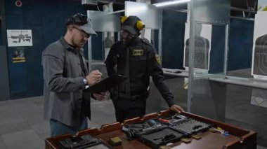 Orta büyüklükte beyaz erkek atış eğitmeni ve Afrika kökenli Amerikalı polis test sonuçlarını gözden geçirip eğitimin başarıyla tamamlanmasını kutluyor ve el sıkışıyor.