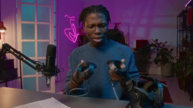 Orta ölçekli genç Afrikalı Afrikalı Amerikalı erkek nüfuzlu stüdyoda oturmuş, el kontrolörleriyle sosyal medyada izleyicilere yeni sanal gerçeklik kulaklığı modelini sunuyor.