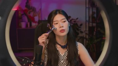 Genç Koreli kadın güzellik blogcusunun canlı yayında yüz masajı silindirini test ederken, yanakları ve çene hattını şekillendirirken, izleyicilere cilt bakımı prosedürlerini anlatırken orta ışıkta orta ölçekli bir çekim.