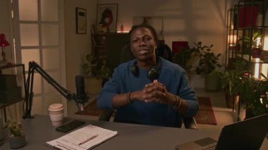 Sosyal medyada canlı yayına başlamadan önce fotoğraf makinesine bakıp gülümseyen, masabaşında podcaster pozu veren neşeli, kendinden emin genç Afrikalı Amerikalı erkek podcaster portresi.