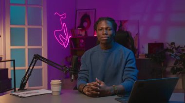 Mavi süveterli, gülümseyen siyah bir blog yazarının orta boy portresi neon ışıklı stüdyoda, dizüstü bilgisayarıyla, mikrofonuyla, akıllı telefonuyla, içerik planıyla, kameraya özgüvenle bakarken.