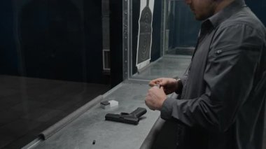 Gri gömlekli ve beysbol şapkalı beyaz bir adam atış poligonunda duruyor, şarjöre mermi dolduruyor, tabancayı yerleştiriyor ve tek başına atış talimi yaparken hedefe ateş ediyor.