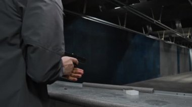 Gri ceketli, beysbol şapkalı ve koruyucu kulaklıklı, kurşunları silaha sokan, ateş eden, hedef alan, hedef alan, hedef tahtasından çıkan genç bir erkek.