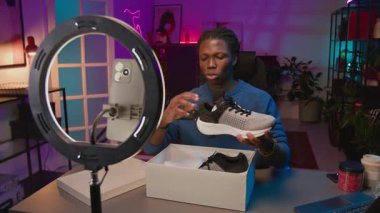 Orta karar Afro-Amerikan erkek blog yazarı örgülü saçlarıyla stüdyoda akıllı telefonuyla halka ışığının önünde oturuyor, sosyal medyada abonelere yeni spor ayakkabıları satıyor.