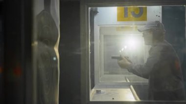 Kulaklık takan, yarı otomatik tabancaya kurşun dolduran, hedef alan ve tetiği çeken, bariyerlere ateş ederken camdan çekilen odaklanmış genç bir adamın arka ışıklandırması.